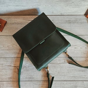May include: A dark green leather backpack with a rectangular flap closure, a main compartment, and adjustable shoulder straps attached with metal hardware. The bag features a smooth finish and clean stitching along the edges of the flap and body.