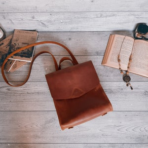 Op de afbeelding: Een middelbruine leren rugzak met een overslagklep met een driehoekig stikseldetail, een handvat aan de bovenkant en verstelbare schouderbanden. De tas heeft een gladde matte afwerking en een gestructureerde rechthoekige vorm met afgeronde hoeken.