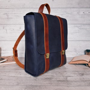 Pode incluir: Uma mochila de couro azul-marinho com uma alça superior e duas tiras verticais de couro castanho com fivelas de metal em tom de latão. A mochila tem alças ajustáveis e um fecho de aba, apresentando um acabamento liso e mate.