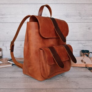 Op de afbeelding: Een beige leren rugzak met een bovenflap en een voorvak, beide vastgezet met donkerbruine leren riemen. De rugzak is voorzien van een handvat aan de bovenkant en verstelbare schouderbanden, met een gladde, matte afwerking over het geheel.