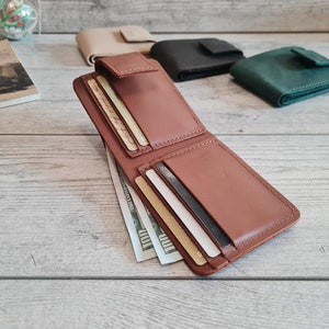 Pu&ograve; includere: Portafoglio in pelle marrone aperto con pi&ugrave; fessure per carte e uno scomparto per contanti contenente diverse banconote da cento dollari. Altri portafogli chiusi beige, neri e verdi sono visibili sullo sfondo.