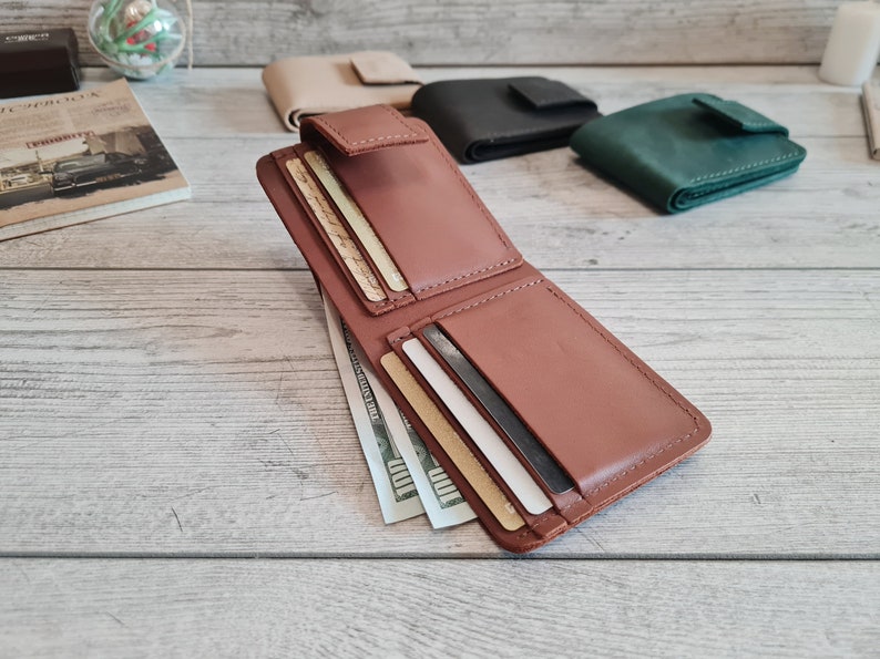Pu&ograve; includere: Portafoglio in pelle marrone aperto con pi&ugrave; fessure per carte e uno scomparto per contanti contenente diverse banconote da cento dollari. Altri portafogli chiusi beige, neri e verdi sono visibili sullo sfondo.