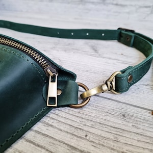 Puede incluir: Una ri&ntilde;onera de cuero verde oscuro con cierre de cremallera met&aacute;lico en tono lat&oacute;n y una correa ajustable a juego sujeta con un mosquet&oacute;n y una anilla en D en tono lat&oacute;n. El bolso presenta costuras visibles en los bordes y una superficie lisa.