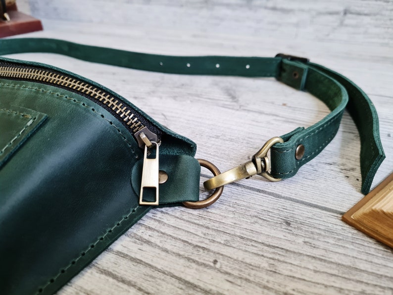 Puede incluir: Una ri&ntilde;onera de cuero verde oscuro con cierre de cremallera met&aacute;lico en tono lat&oacute;n y una correa ajustable a juego sujeta con un mosquet&oacute;n y una anilla en D en tono lat&oacute;n. El bolso presenta costuras visibles en los bordes y una superficie lisa.