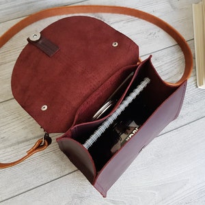 K&ouml;nnte beinhalten: Burgunderfarbene Leder-Umh&auml;ngetasche mit einem langen, hellbraunen Riemen. Die Tasche ist ge&ouml;ffnet und zeigt ein Notizbuch, einen Stift und andere Gegenst&auml;nde. Sie hat einen Klappverschluss und ein strukturiertes Design.
