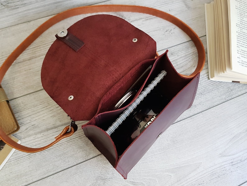 K&ouml;nnte beinhalten: Burgunderfarbene Leder-Umh&auml;ngetasche mit einem langen, hellbraunen Riemen. Die Tasche ist ge&ouml;ffnet und zeigt ein Notizbuch, einen Stift und andere Gegenst&auml;nde. Sie hat einen Klappverschluss und ein strukturiertes Design.