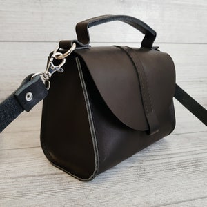 K&ouml;nnte beinhalten: Dunkelbraune Lederhandtasche mit einem oberen Griff und einem abnehmbaren Schulterriemen. Die Tasche hat einen Klappenverschluss und silberfarbene Beschl&auml;ge. Die Tasche hat eine rechteckige Form und scheint handgefertigt zu sein.