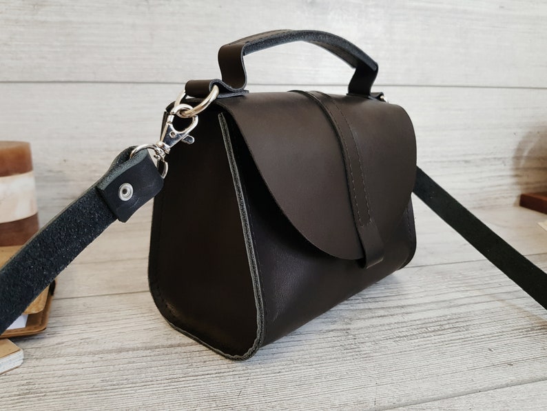 K&ouml;nnte beinhalten: Dunkelbraune Lederhandtasche mit einem oberen Griff und einem abnehmbaren Schulterriemen. Die Tasche hat einen Klappenverschluss und silberfarbene Beschl&auml;ge. Die Tasche hat eine rechteckige Form und scheint handgefertigt zu sein.
