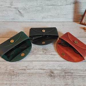 May include: Three leather eyeglass cases in green, black, and reddish-brown, each with a gold snap closure. The cases are open, revealing dark sunglasses inside. The cases are arranged on a light wood-grain surface.