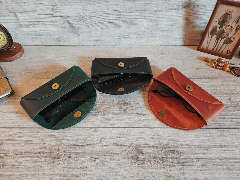 May include: Three leather eyeglass cases in green, black, and reddish-brown, each with a gold snap closure. The cases are open, revealing dark sunglasses inside. The cases are arranged on a light wood-grain surface.