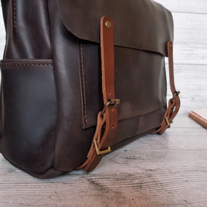 May include: A dark brown leather backpack featuring a large front flap secured by two lighter tan leather straps with antique brass-toned metal buckles. The bag has visible side pockets and reinforced stitching along the edges of the main compartment.