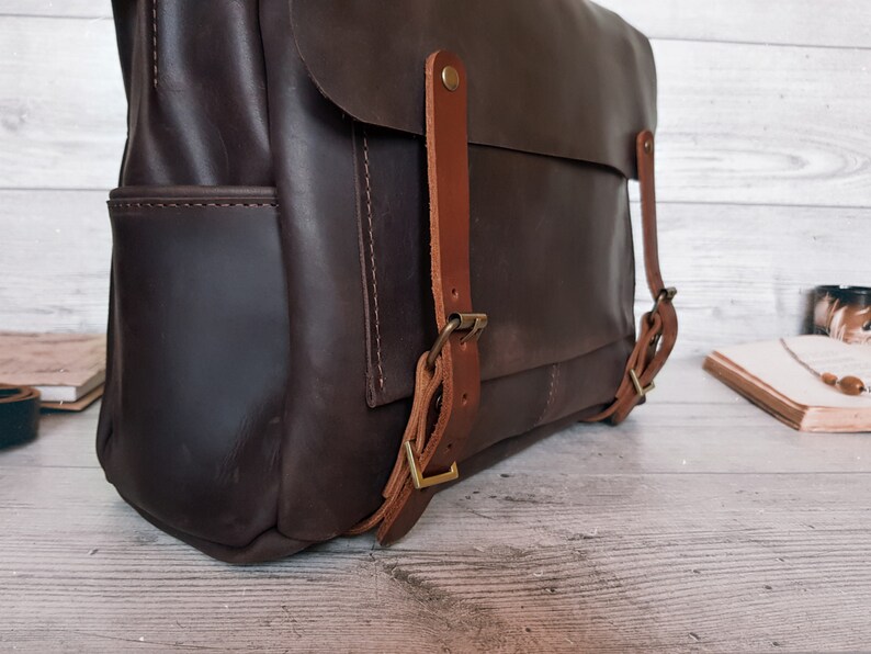 May include: A dark brown leather backpack featuring a large front flap secured by two lighter tan leather straps with antique brass-toned metal buckles. The bag has visible side pockets and reinforced stitching along the edges of the main compartment.