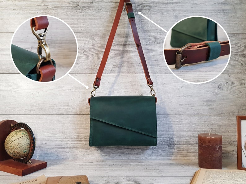 May include: A dark green leather crossbody bag with a diagonal front flap and a long, adjustable brown leather shoulder strap. The bag features antique brass-toned metal hardware, including a swivel lobster clasp and a buckle for strap adjustment.