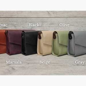 May include: Six rectangular crossbody bags with flap closures, arranged in a row. The colors from left to right are labeled as Cognac, Marsala, Black, Beige, Olive, and Gray. Each bag features a dark brown side tab and a smooth, solid finish.