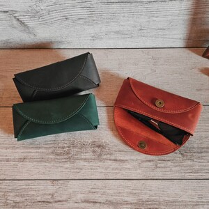 May include: Three leather eyeglass cases in shades of black, green, and red-orange. The cases are closed and open, revealing a pair of sunglasses. The cases are on a light wood surface.