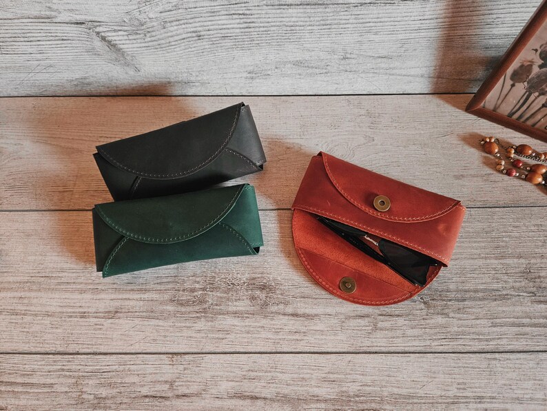 May include: Three leather eyeglass cases in shades of black, green, and red-orange. The cases are closed and open, revealing a pair of sunglasses. The cases are on a light wood surface.