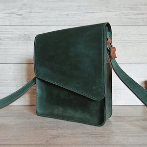 May include: A dark green crossbody bag with a rectangular silhouette and a fold-over flap closure. The bag features a matching green adjustable shoulder strap attached with brown leather accents on the sides, showcasing a smooth matte finish.