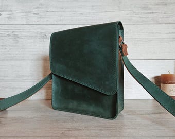 May include: A dark green crossbody bag with a rectangular silhouette and a fold-over flap closure. The bag features a matching green adjustable shoulder strap attached with brown leather accents on the sides, showcasing a smooth matte finish.