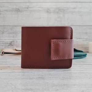 Pu&ograve; includere: Un portafoglio in pelle marrone con una chiusura a patta marrone rosato a contrasto. Il portafoglio &egrave; rettangolare con angoli arrotondati e cuciture visibili. Lo sfondo &egrave; una superficie di legno chiaro.