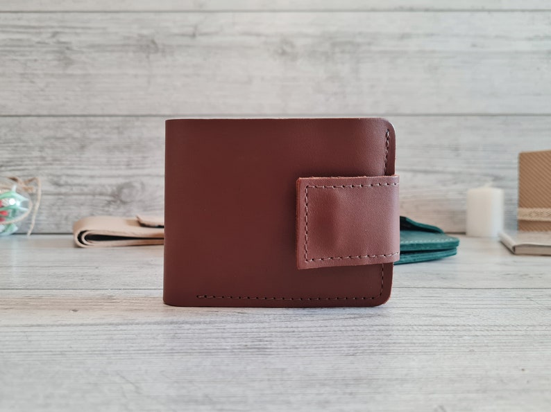 Pu&ograve; includere: Un portafoglio in pelle marrone con una chiusura a patta marrone rosato a contrasto. Il portafoglio &egrave; rettangolare con angoli arrotondati e cuciture visibili. Lo sfondo &egrave; una superficie di legno chiaro.