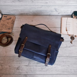 May include: A dark blue leather messenger bag featuring a top handle and a front flap secured by two brown leather straps with brass-toned metal buckles. The bag includes a detachable brown leather shoulder strap coiled to the left side.
