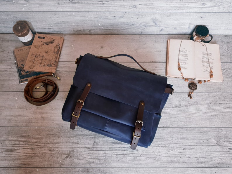 May include: A dark blue leather messenger bag featuring a top handle and a front flap secured by two brown leather straps with brass-toned metal buckles. The bag includes a detachable brown leather shoulder strap coiled to the left side.