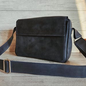 May include: A dark charcoal gray rectangular crossbody bag featuring a smooth flap closure, a wide adjustable shoulder strap with a metallic gold-toned buckle, and visible tonal stitching along the edges of the structured leather-like material.
