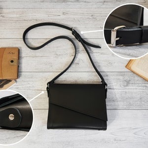 May include: A black crossbody bag with a smooth finish and a flap closure featuring an asymmetrical diagonal cut. The bag includes a long, adjustable shoulder strap with a silver-toned metal buckle and side attachments secured with silver-toned rivets.