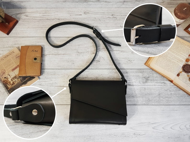 May include: A black crossbody bag with a smooth finish and a flap closure featuring an asymmetrical diagonal cut. The bag includes a long, adjustable shoulder strap with a silver-toned metal buckle and side attachments secured with silver-toned rivets.