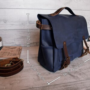 May include: A blue leather satchel with a top handle and brown leather straps. The bag measures 11 inches in height, 5.9 inches in depth, and 16.5 inches in width. It includes a detachable brown leather shoulder strap with metal hardware.