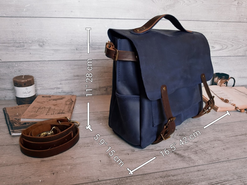 May include: A blue leather satchel with a top handle and brown leather straps. The bag measures 11 inches in height, 5.9 inches in depth, and 16.5 inches in width. It includes a detachable brown leather shoulder strap with metal hardware.