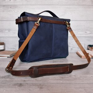 May include: A navy blue leather messenger bag featuring a top handle and a detachable, adjustable brown leather shoulder strap with a padded section. The bag includes brass-toned metal hardware, including swivel clips and a buckle for strap length.
