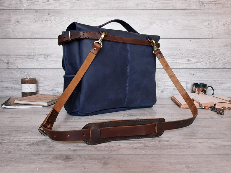 May include: A navy blue leather messenger bag featuring a top handle and a detachable, adjustable brown leather shoulder strap with a padded section. The bag includes brass-toned metal hardware, including swivel clips and a buckle for strap length.