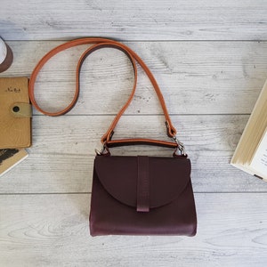 K&ouml;nnte beinhalten: Burgunderfarbene Leder-Umh&auml;ngetasche mit hellbraunem Riemen. Die Tasche hat einen Klappenverschluss und einen kleinen Griff. Der Riemen ist verstellbar. Die Tasche liegt auf einer hellen Holzoberfl&auml;che mit einem Buch, einem Notizbuch und einer Kerze.