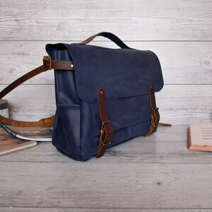 May include: A navy blue leather messenger bag featuring a top handle, a fold-over flap with two brown leather straps, and brass-toned metal buckles. The bag includes side pockets and an adjustable brown leather shoulder strap with metal hardware.