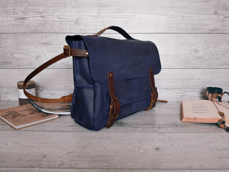 May include: A navy blue leather messenger bag featuring a top handle, a fold-over flap with two brown leather straps, and brass-toned metal buckles. The bag includes side pockets and an adjustable brown leather shoulder strap with metal hardware.