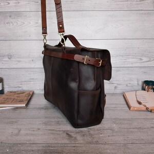 May include: A dark brown leather messenger bag featuring a top handle, a detachable shoulder strap with metal clips, and a horizontal strap across the front secured with a brass-toned metal buckle. The bag has a rectangular shape and side pockets.