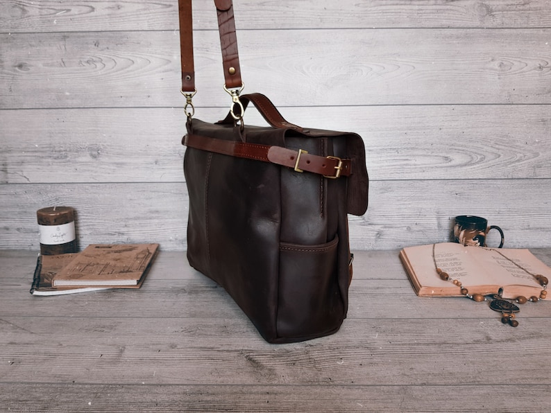 May include: A dark brown leather messenger bag featuring a top handle, a detachable shoulder strap with metal clips, and a horizontal strap across the front secured with a brass-toned metal buckle. The bag has a rectangular shape and side pockets.
