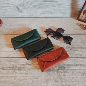 May include: Three leather eyeglass cases in green, black, and red-orange are displayed on a wooden surface, alongside two pairs of sunglasses. A small globe and framed artwork are visible in the background.