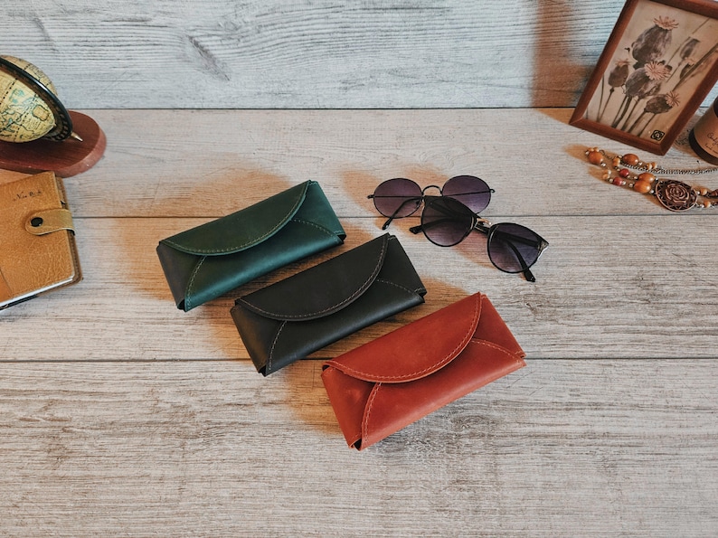 May include: Three leather eyeglass cases in green, black, and red-orange are displayed on a wooden surface, alongside two pairs of sunglasses. A small globe and framed artwork are visible in the background.