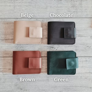 Pu&ograve; includere: Quattro portafogli in pelle beige, cioccolato, marrone e verde. Ogni portafoglio ha una forma rettangolare con chiusura a patta quadrata. I portafogli sono esposti su una superficie chiara con venature del legno.