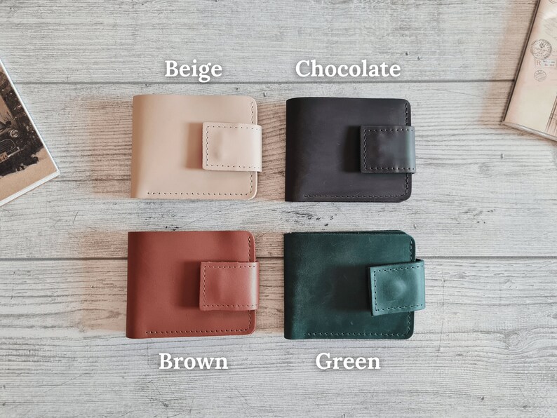 Pu&ograve; includere: Quattro portafogli in pelle beige, cioccolato, marrone e verde. Ogni portafoglio ha una forma rettangolare con chiusura a patta quadrata. I portafogli sono esposti su una superficie chiara con venature del legno.