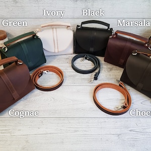 K&ouml;nnte beinhalten: Eine Auswahl an Lederhandtaschen in verschiedenen Farben: Gr&uuml;n, Elfenbein, Schwarz, Marsala, Cognac und Schokolade. Jede Tasche hat einen oberen Griff und einen abnehmbaren Schulterriemen. Die Taschen werden auf einer hellen Holzoberfl&auml;che pr&auml;sentiert.