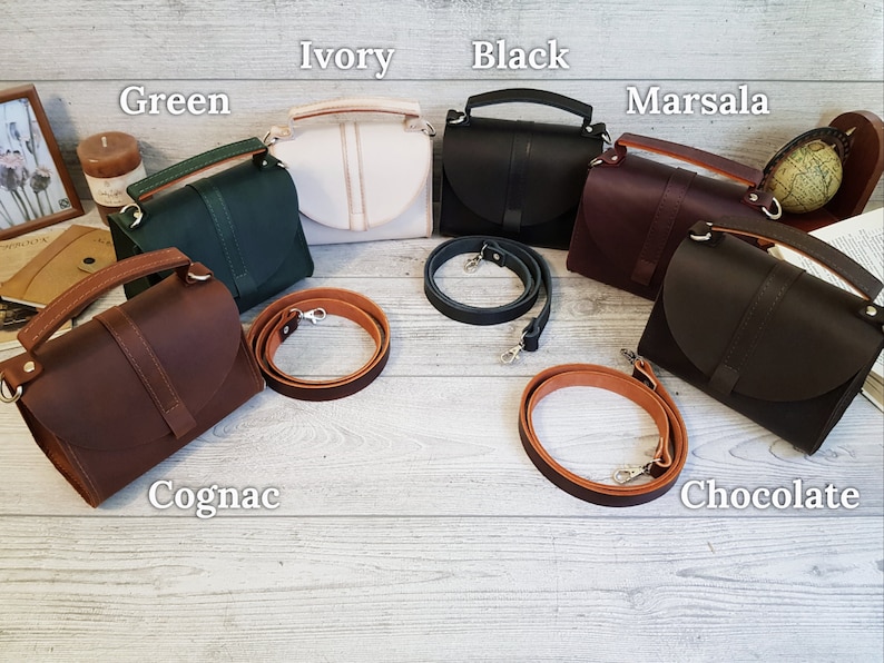 K&ouml;nnte beinhalten: Eine Auswahl an Lederhandtaschen in verschiedenen Farben: Gr&uuml;n, Elfenbein, Schwarz, Marsala, Cognac und Schokolade. Jede Tasche hat einen oberen Griff und einen abnehmbaren Schulterriemen. Die Taschen werden auf einer hellen Holzoberfl&auml;che pr&auml;sentiert.