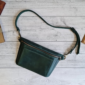 Puede incluir: Un bolso bandolera de cuero verde oscuro con un compartimento principal rectangular con cierre de cremallera superior, un bolsillo frontal y una correa ajustable para el hombro sujeta con clips de metal en tono lat&oacute;n antiguo.