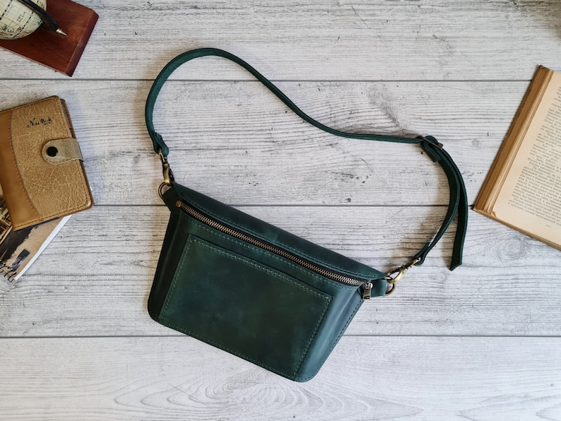 Puede incluir: Un bolso bandolera de cuero verde oscuro con un compartimento principal rectangular con cierre de cremallera superior, un bolsillo frontal y una correa ajustable para el hombro sujeta con clips de metal en tono lat&oacute;n antiguo.