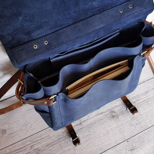 May include: A navy blue leather messenger bag with a large flap, featuring multiple interior compartments and pockets. The bag includes brown leather straps with brass-toned metal buckles and rivets, and it is shown open with books inside.