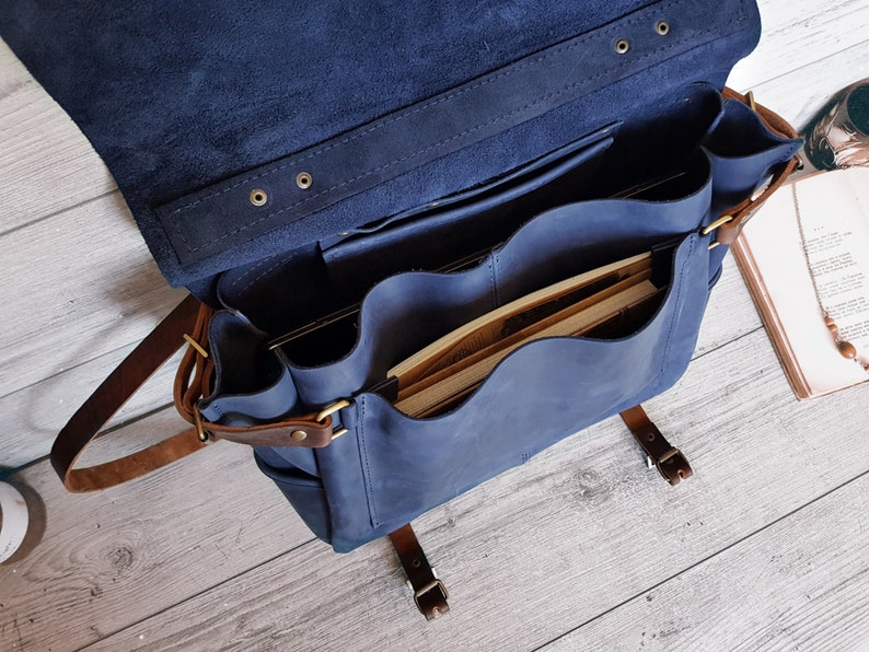May include: A navy blue leather messenger bag with a large flap, featuring multiple interior compartments and pockets. The bag includes brown leather straps with brass-toned metal buckles and rivets, and it is shown open with books inside.