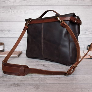 May include: A dark brown leather messenger bag featuring a top handle and an adjustable shoulder strap with a padded section. The bag has a fold-over flap, brass-toned metal hardware, and visible stitching along the front panel and strap attachments.