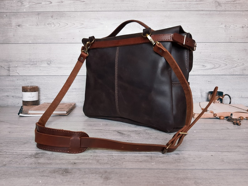 May include: A dark brown leather messenger bag featuring a top handle and an adjustable shoulder strap with a padded section. The bag has a fold-over flap, brass-toned metal hardware, and visible stitching along the front panel and strap attachments.